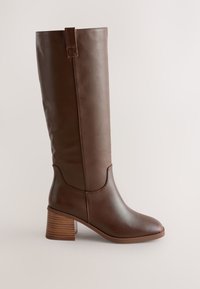 Botte en cuir marron montant jusqu'au genou avec talon bloc et languette de tirage sur fond clair uni, vue de côté.