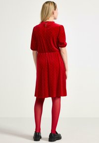 Czerwona sukienka z weluru z krótkimi bufiastymi rękawami, marszczoną talią i małym guzikiem z tyłu. Połączona z czerwonymi rajstopami i czarnymi butami.