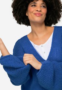 Blauwe gebreide cardigan met een losse pasvorm en diepe V-hals, textuur garen, oversized mouwen, gedragen over een wit overhemd met knopen.