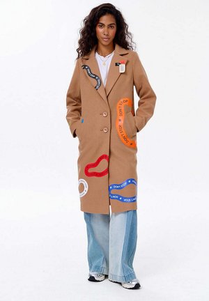 Žena v béžovém kabátu s barevnými nášivkami "NEZASTAVUJ", v světl�ých modrých širokých džínách, v bílých teniskách, stojící s rukama v kapsách.