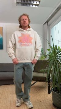 Kremowa bluza z kapturem z graficznym nadrukiem pomarańczowego słońca, zestawiona z jasnozielonymi spodniami i szarymi tenisówkami. Wnętrze z roślinami.