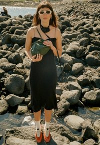 Juoda be rankovių suknelė, žalia kryžminė rankinė, baltos saulės akys, raudoni batai su baltais kojinėlėmis. Stovi ant akmenuoto reljefo netoli vandens.