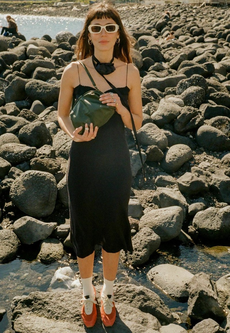 Juoda be rankovių suknelė, žalia kryžminė rankinė, baltos saulės akys, raudoni batai su baltais kojinėlėmis. Stovi ant akmenuoto reljefo netoli vandens.