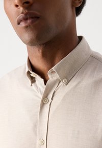 Μπεζ πουκάμισο με κολάρο και λεία υφή, με σχέδιο με κουμπιά και λεπτές διακοσμητικές ραφές στο κολάρο και την πατιλέτα.