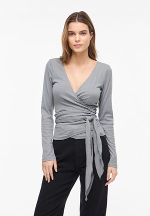 Nő hosszú ujjú szürke átkötős felsőben, amely deréknál meg van kötve, és fekete nadrágban, egyszerű fehér háttér előtt áll.