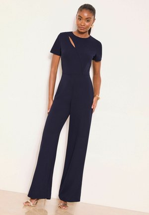 Woman wearing a navy blue short-sleeve jumpsuit with wide-leg pants and a small chest cutout, paired with gold high-heeled sandals.
