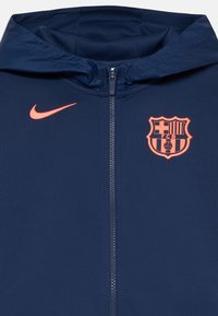 Marineblauwe Nike-jack met een rits, voorzien van een oranje swoosh-logo en het FC Barcelona-embleem op de borst, met een gladde stoftextuur.