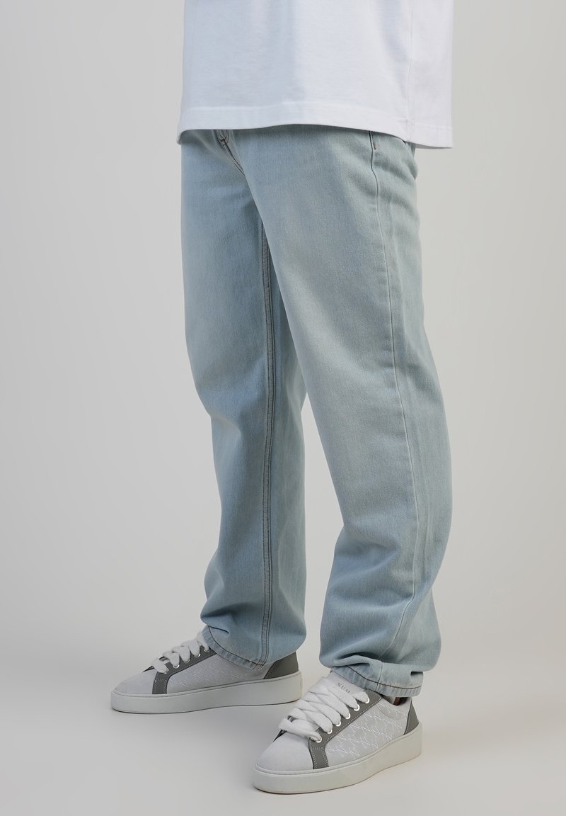Person, die hellblaue Jeans und weiße Sneakers mit grauen Akzenten trägt, vor einem schlichten hellen Hintergrund stehend.