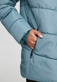 Tirkīza krāsas pūkains mētels ar rāvējslēdzēju kabatu. Iezīmējas ar quilted faktūru un ribotām piedurknēm. Izgatavots no viegla, ūdensizturīga materiāla.