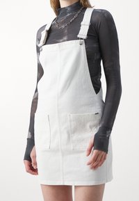 Biały ogrodniczy sukienka z denimu z regulowanymi ramiączkami, z dwoma przednimi kieszeniami z detalami w formie oczek. Gładka tekstura i dopasowany krój.