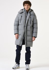 Abrigo acolchado gris claro con capucha, costuras detalladas y múltiples bolsillos con cremallera; combinado con pantalones oscuros y zapatillas deportivas.