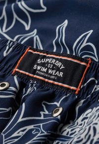 Marine zwemkleding met een wit bloemenpatroon, elastische tailleband en een gemarkeerd zwart label met oranje accenten. Zilveren drukknoopdetails.