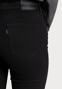 Jeans de mezclilla negra con cintura alta, que presentan un diseño clásico de bolsillo trasero y detalles de costura. Parche de marca de cuero en la cintura.