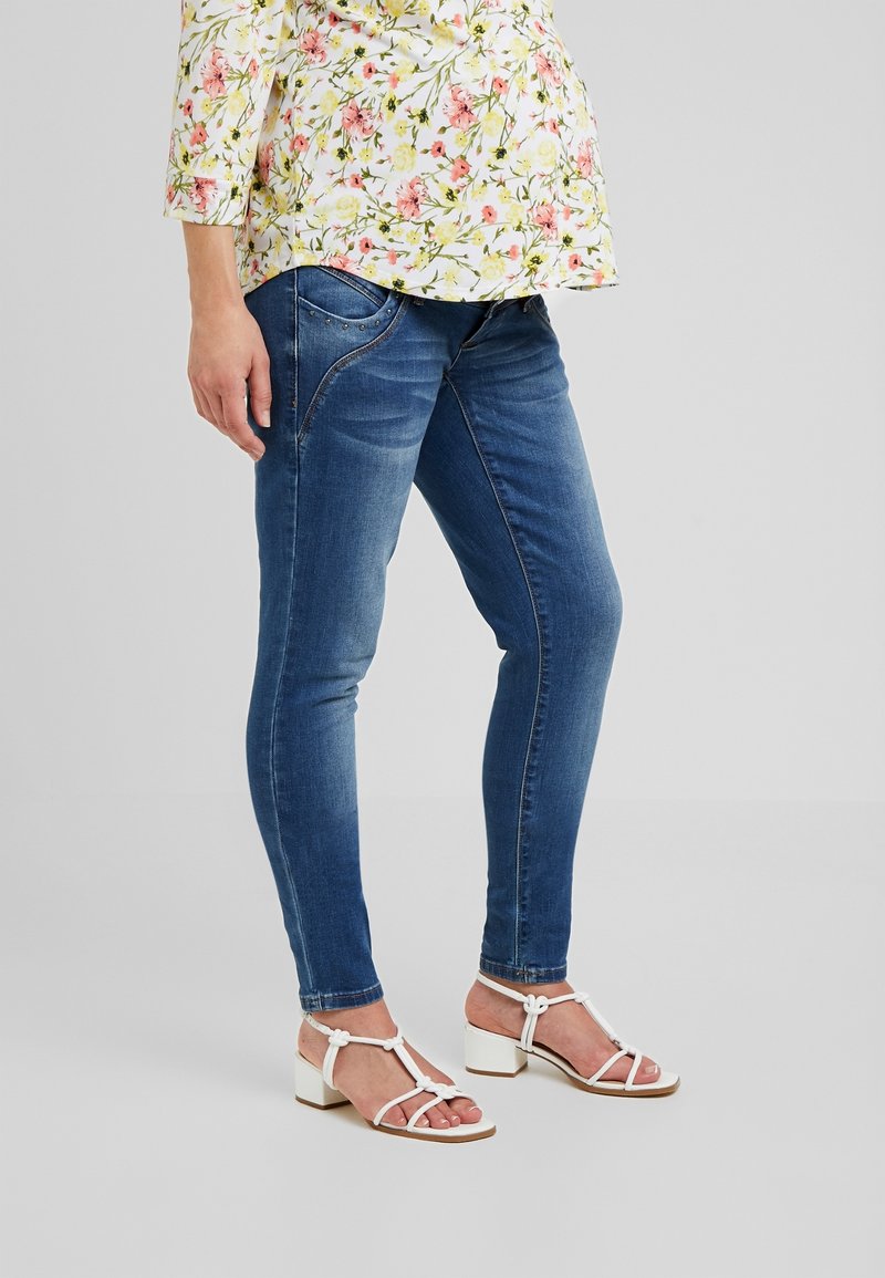 Blue denim skinny jeans with a fitted shape, paired with white strappy sandals featuring a low block heel. Floral patterned top.