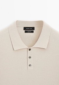 Beige bomuldspolo med en ribbet struktur, forsynet med en tre-knaps stolpelukning og en klassisk krave. Mærkeetiket synlig indvendigt.