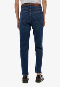 Jeans de mezclilla de un azul oscuro con una cintura alta, que cuentan con dos bolsillos traseros. Combinados con un cinturón trenzado marrón y calzado negro.