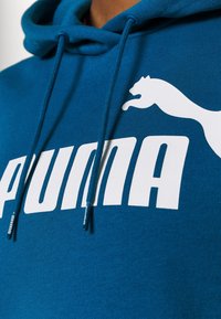Modrá mikina s kapucí vyrobená z měkké tkaniny s velkým bílým logem „PUMA“ a siluetou kočky, s tkaničkami a žebrovanými detaily.