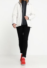 Persoon in een witte hoodiejack, zwarte broek, grijze ritsvest en felrode sneakers die vooruit loopt op een eenvoudige witte achtergrond.