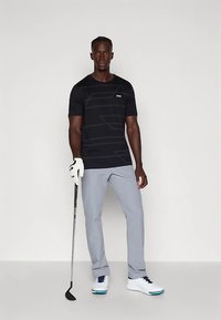 Black short-sleeve shirt with subtle striped pattern, gray pants, white golf shoes, holding a golf club, posing on a neutral background.
