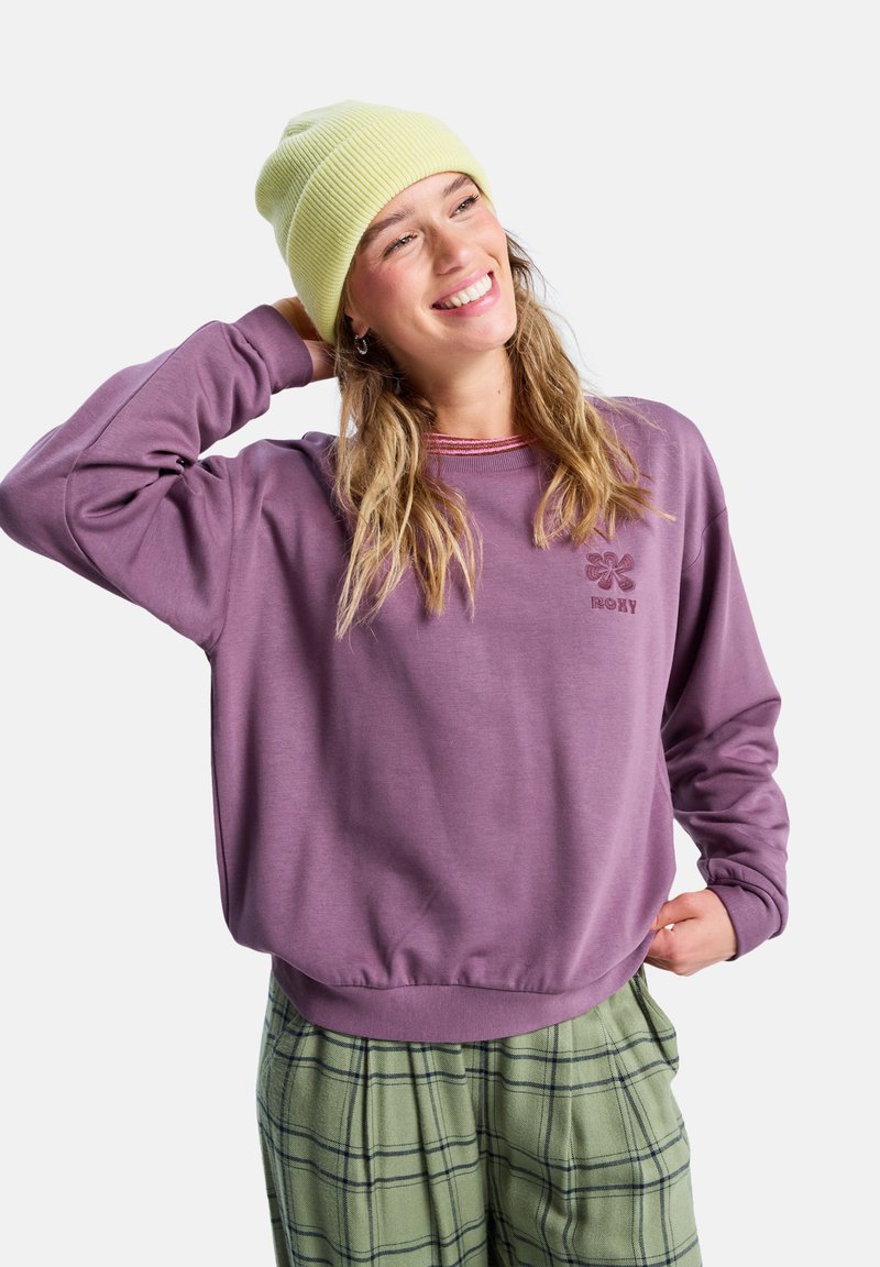Sudadera de cuello redondo en suave color púrpura con puños de canalé. Presenta un logo floral en púrpura más oscuro. Combinada con pantalones de cuadros verdes.