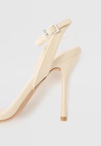 Beige høyhælte sandaler med et elegant design, som har justerbar ankelrem og spiss tå. Glatt tekstur med en blank overflate.