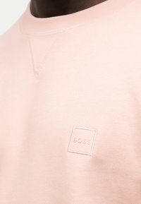 Κοντινό ενός ατόμου που φοράει ανοιχτό ροζ φούτερ με μικρό ανάγλυφο λογότυπο BOSS στο στήθος.