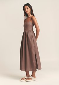 Brun, lång, ärmlös klänning med figursydd överdel, rynkad kjol och slits i sidan. Har tunna band och slät textur. Bärs med beiga sandaler.