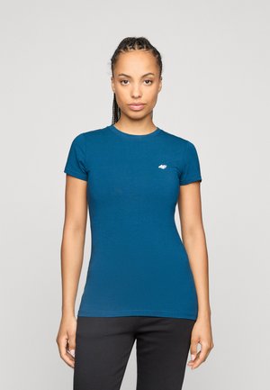 Vrouw met gevlochten haar, een aansluitend blauwe korte mouw T-shirt en zwarte broek dragend, staand tegen een effen lichte achtergrond.