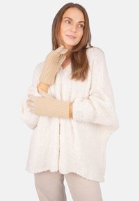 Frau in einem cremefarbenen grob gestrickten Pullover und beigen Handschuhen, die seitlich schaut und sanft lächelt, mit erhobenen Händen nahe Gesicht und Brust.