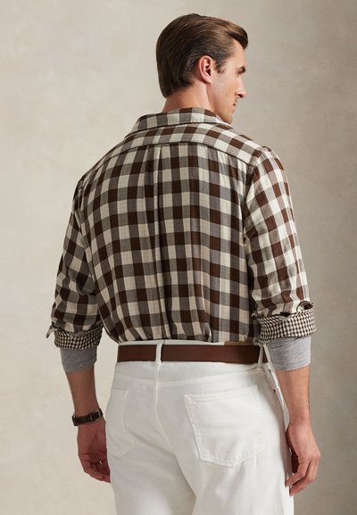 Homem vestindo camisa xadrez marrom e branca sobre uma camiseta cinza de manga comprida, calças brancas e cinto castanho, visto de costas contra um fundo neutro.