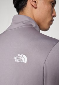 Γκρι πουλόβερ με υψηλ ό γιακά, κατασκευασμένο από λείο, υφή ύφασμα· διαθέτει το λογότυπο The North Face σε λευκό στην πλάτη.