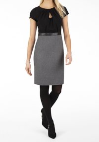 Robe noire sans manches avec un détail en forme de trou de serrure, jupe crayon en laine grise, coupe ajustée, ceinture en cuir noire, portée avec des collants noirs et des talons.
