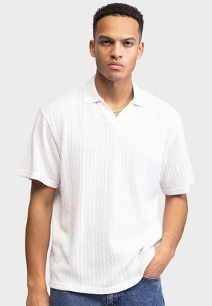 Νέος άνδρας με κοντά σγουρά μαλλιά, φοράει ένα λευκό υφασμάτινο κοντομάνικο πουκάμισο και μπλε τζιν, στέκεται με το ένα χέρι στην τσέπη.