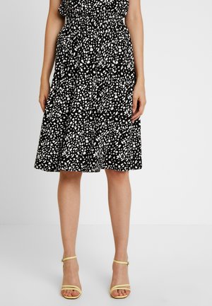 Jupe noire avec imprimé abstrait blanc, taille élastique, longueur genou, design fluide. Associée avec des sandales jaunes à lanières.