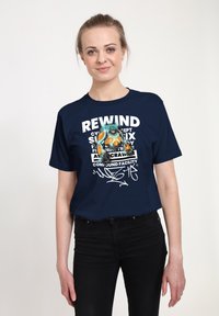 Žena s blond vlasy na sobě má námořnickou modrou tričko s robotickým armovaným postavou a bílým textem s nápisem "REWIND" a dalšími stylizovanými slovy.