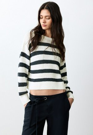 Giovane donna con lunghi capelli scuri che indossa un maglione corto bianco con strisce grigie e pantaloni neri, con le mani nelle tasche, guarda verso il basso.