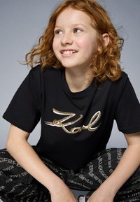 Μαύρο μπλουζάκι με μεγάλη, λαμπερή λογότυπο "Karl" σε χρυσό, συνδυασμένο με μαύρο παντελόνι που φέρει επαναλαμβανόμενη λευκή γραφή. Ίσια ύφασμα και άνετη εφαρμογή.