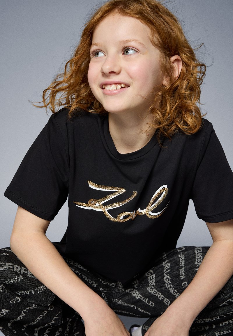 Μαύρο μπλουζάκι με μεγάλη, λαμπερή λογότυπο "Karl" σε χρυσό, συνδυασμένο με μαύρο παντελόνι που φέρει επαναλαμβανόμενη λευκή γραφή. Ίσια ύφασμα και άνετη εφαρμογή.