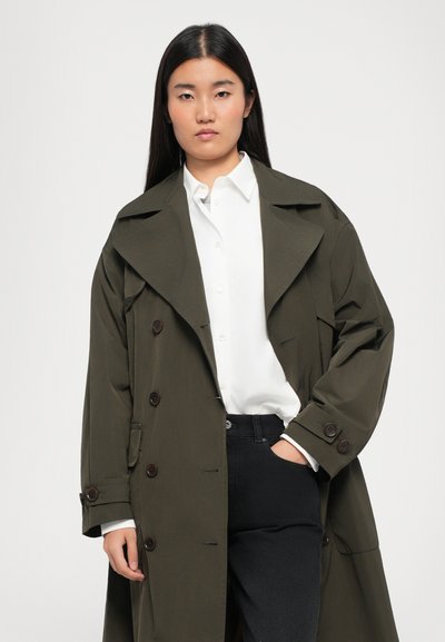 Olivgrüner, doppelt brechender Trenchcoat mit großem Kragen, Knopfdetails und hochgekrempelten Ärmeln, getragen über einem weißen Hemd mit Kragen und schwarzen Hosen.
