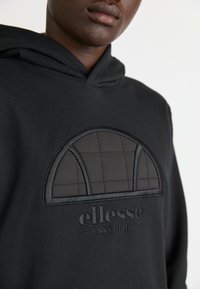 Sudadera negra con capucha que presenta un parche texturizado con un diseño arqueado, un patrón cosido y el logo bordado "ellesse Ski Club" en la parte frontal.