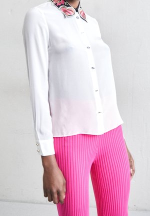 Osoba ubrana w białą bluzkę z kołnierzem ozdobionym kwiatowym haftem i błyszczącymi guzikami, w połączeniu z jasnoróżowymi prążkowanymi spodniami.