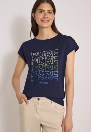 Jeune femme aux longs cheveux bruns souriante, portant un t-shirt bleu marine avec un texte coloré "PURE" et un pantalon beige clair.