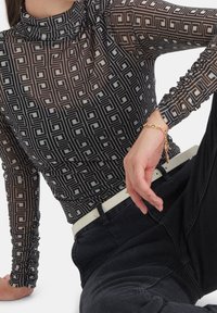 Mustap distintivinen paita, jossa on pitkä hiha ja geometrinen kuvio, korkealla kauluksella ja kerrostetuilla yksityiskohdilla. Yhdistetty mustiin farkkuihin ja valkoiseen vyöhön.