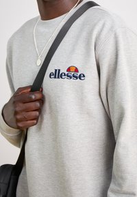 Personne portant un sweat-shirt ellesse gris clair, tenant la sangle noire d'un sac sur l'épaule, et un collier argenté avec un pendentif ovale visible.