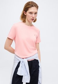 Νεαρή γυναίκα που φοράει ανοιχτό ροζ μπλουζάκι και σκούρο παντελόνι, με ένα γαλάζιο ριγέ πουκάμισο δεμένο γύρω από τη μέση της, μπροστά σε απλό φόντο.