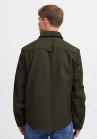 Giacca camicia di lana verde oliva con colletto a punta, maniche lunghe e chiusura con bottoni. Presenta un anello sul retro e cuciture robuste.