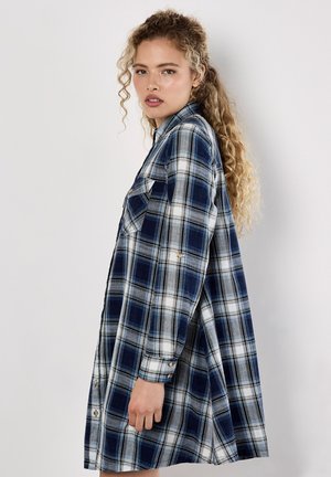 Vrouw met lang krullend haar, gekleed in een blauw-witte geruite overhemdjurk, staande zijwaarts tegen een effen witte achtergrond.