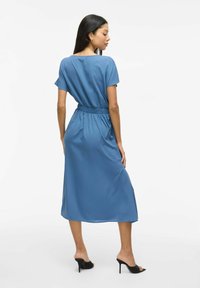 Rochie midi albastră dintr-o țesătură netedă, cu o croială lejeră, mâneci scurte, talie elastică și fustă dreaptă. Pantofi negri cu toc înalt vizibili.