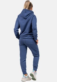 Completo con felpa blu e pantaloni della tuta realizzati in tessuto morbido. Presenta un cappuccio, polsini a costine e una tasca sul retro. Abbinato a sneakers di colore chiaro.