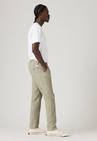 Man with braided hair stands facing left, wearing white t-shirt, beige pants, and white sneakers with black and red details.